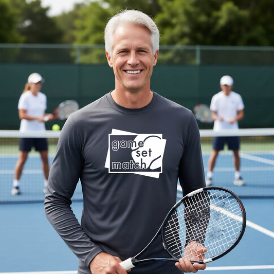 A man on a blue tennis court holds a tennis racket and wears a dark gray long-sleeve T-shirt with a white graphic of two overlapping white and gray squares with the cut out words of ‘GAME SET MATCH” and a tennis ball, printed on the front of the shirt. 