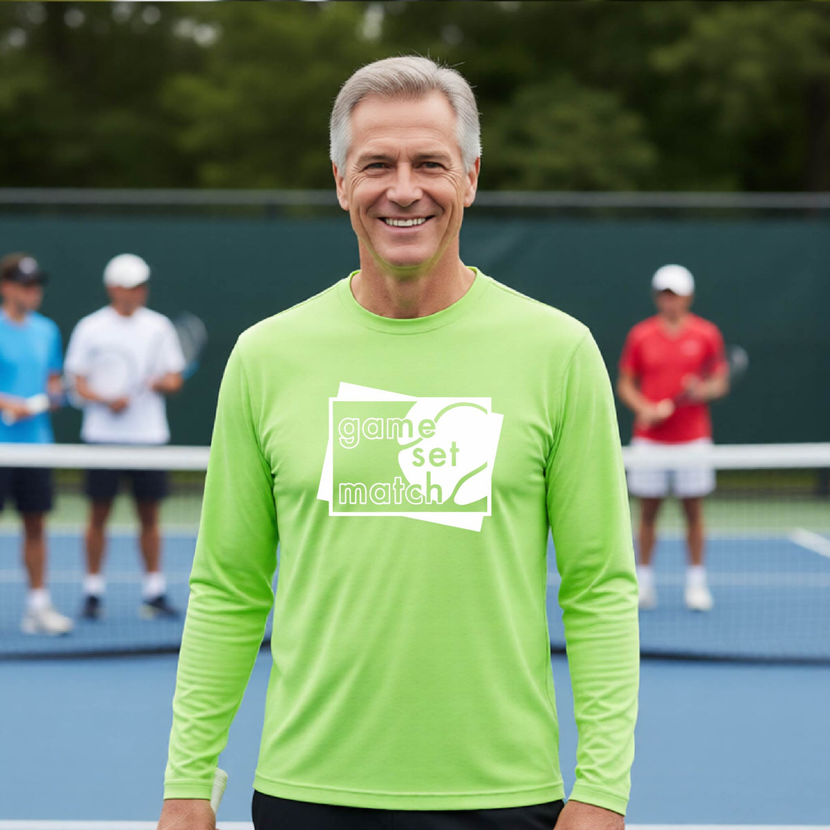 A man on a blue tennis court holds a tennis racket and wears a lime green long-sleeve T-shirt with a white graphic of two overlapping white and gray squares with the cut out words of ‘GAME SET MATCH” and a tennis ball, printed on the front of the shirt. 