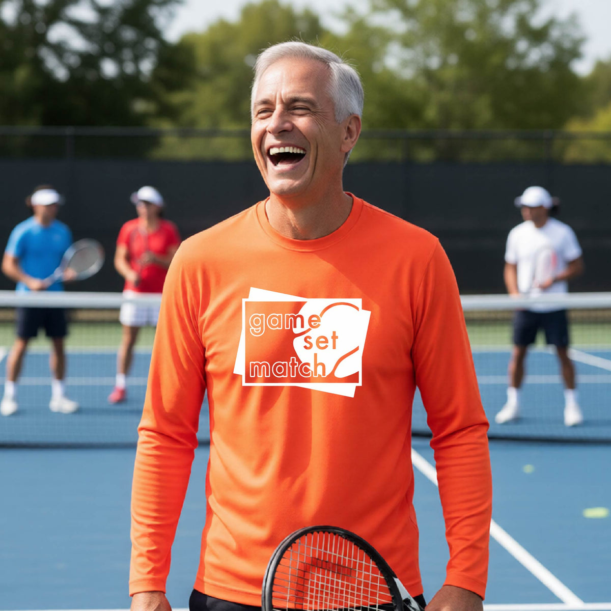A man on a blue tennis court holds a tennis racket and wears an orange long-sleeve T-shirt with a white graphic of two overlapping white and gray squares with the cut out words of ‘GAME SET MATCH” and a tennis ball, printed on the front of the shirt. 