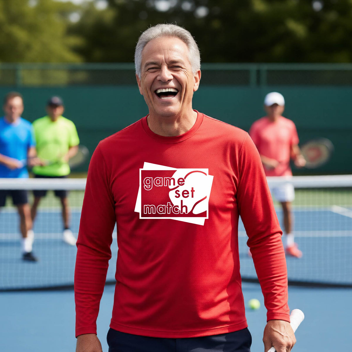 A man on a blue tennis court holds a tennis racket and wears a red long-sleeve T-shirt with a white graphic of two overlapping white and gray squares with the cut out words of ‘GAME SET MATCH” and a tennis ball, printed on the front of the shirt. 