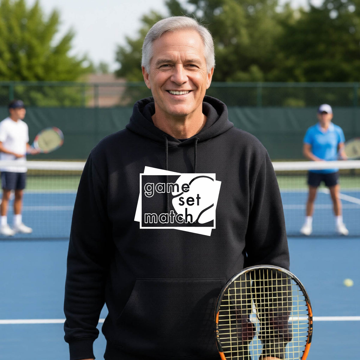 A man on a blue tennis court holds a tennis racket and wears a black unisex hoodie pullover with a white graphic of two overlapping white and gray squares with the cut out words of ‘GAME SET MATCH” and a tennis ball, printed on the front of the shirt. 