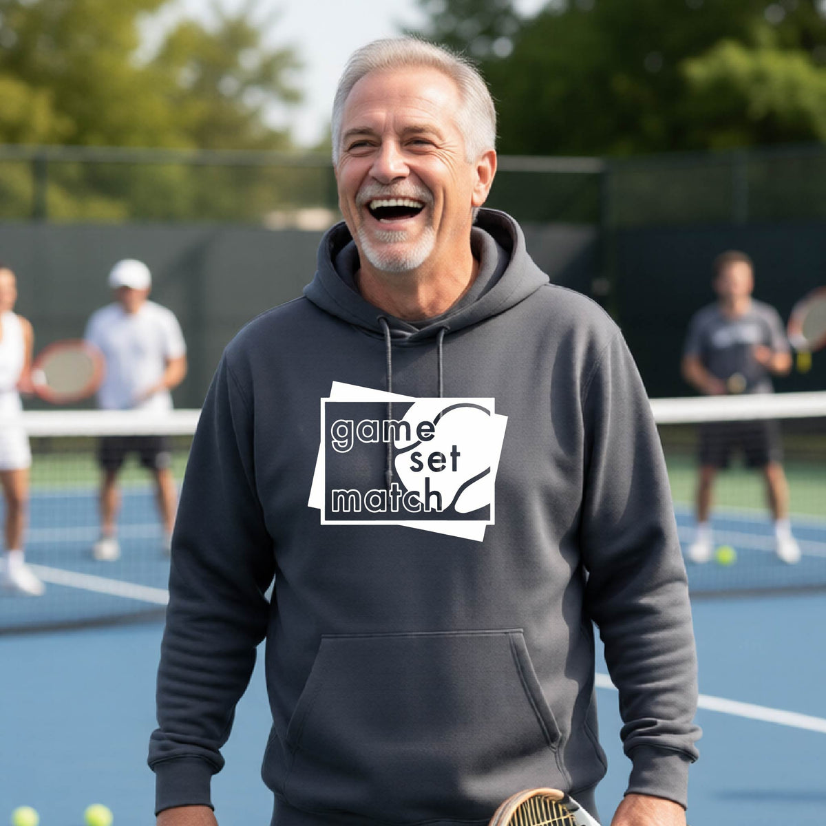 A man on a blue tennis court holds a tennis racket and wears a dark gray unisex hoodie pullover with a white graphic of two overlapping white and gray squares with the cut out words of ‘GAME SET MATCH” and a tennis ball, printed on the front of the shirt. 