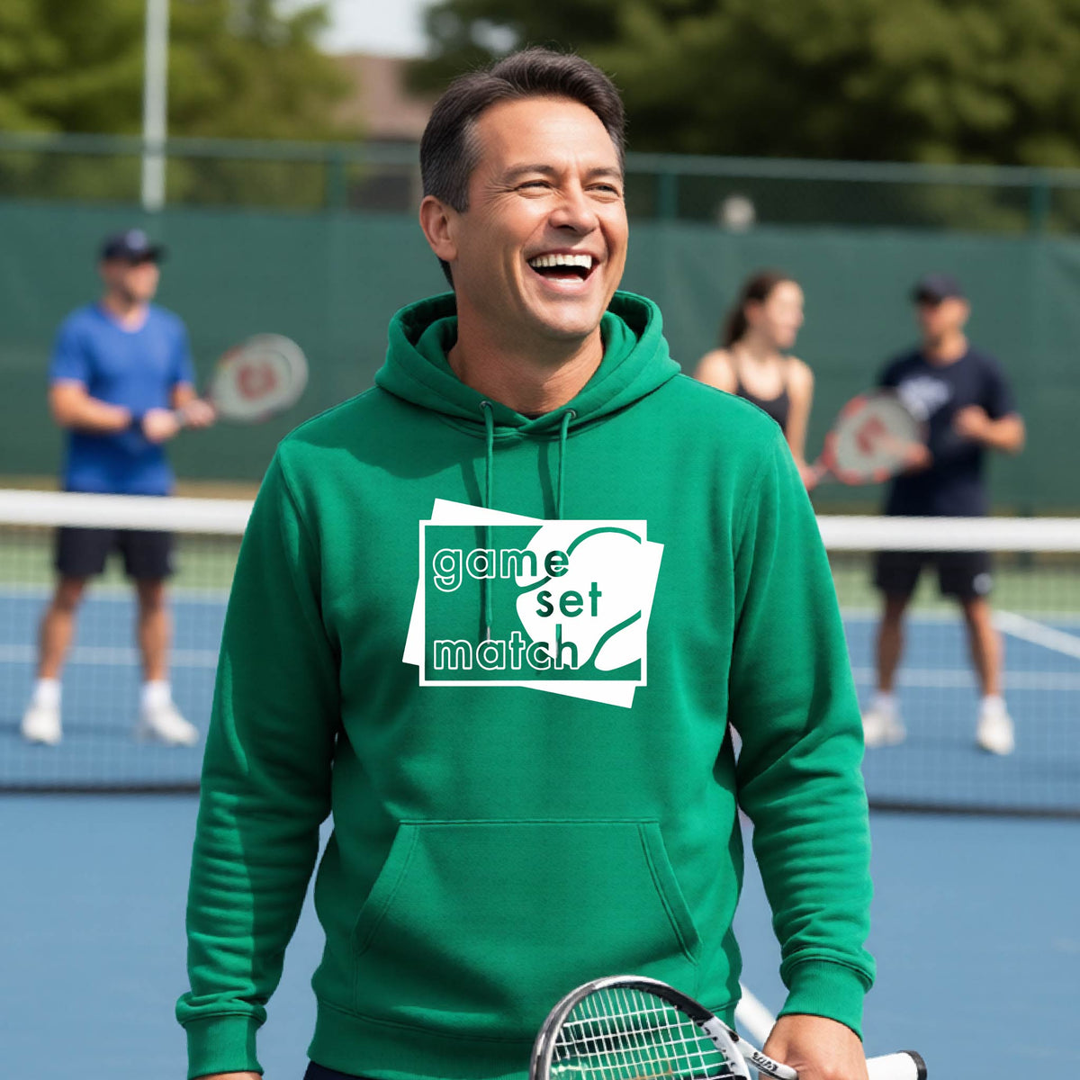 A man on a blue tennis court holds a tennis racket and wears a green unisex hoodie pullover with a white graphic of two overlapping white and gray squares with the cut out words of ‘GAME SET MATCH” and a tennis ball, printed on the front of the shirt. 