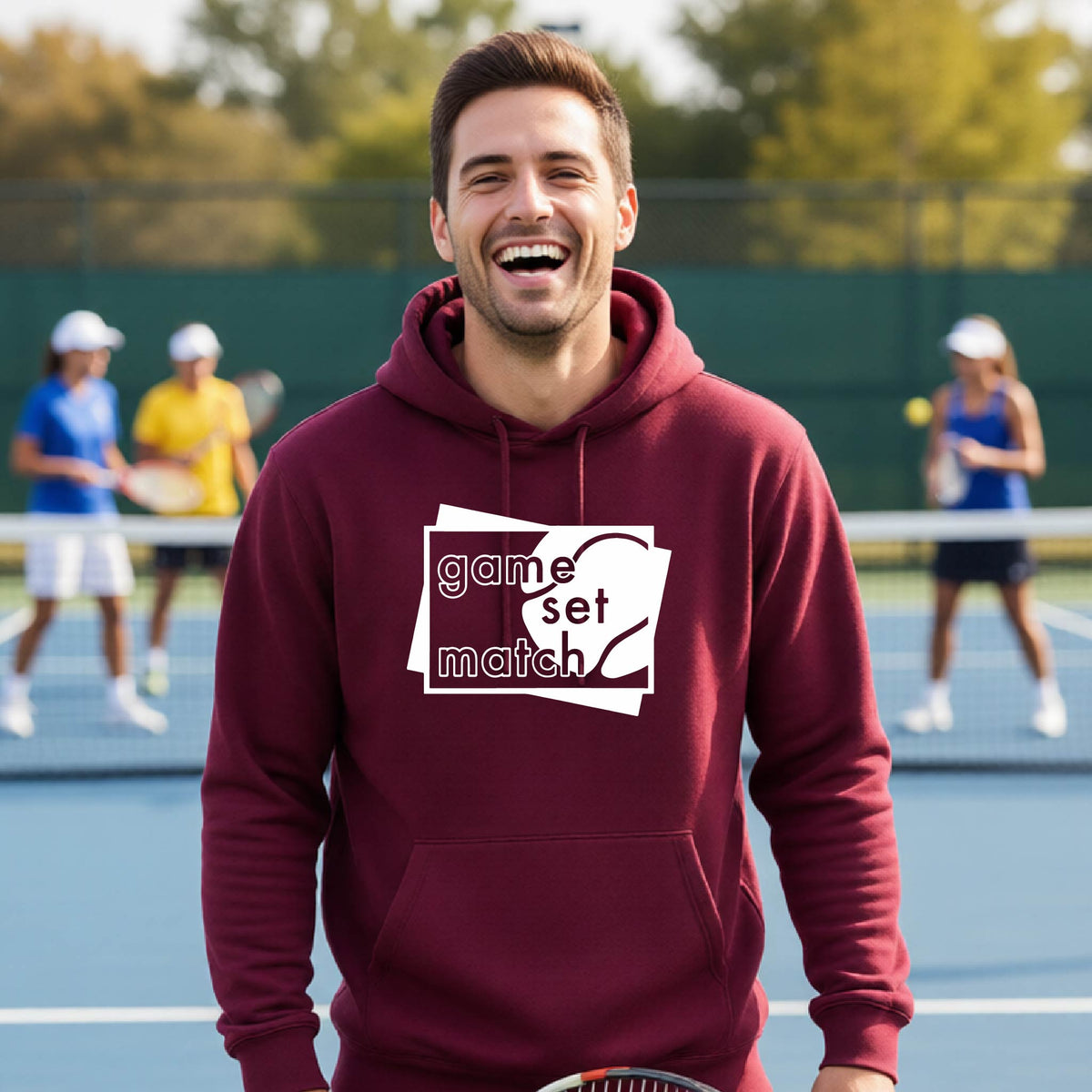 A man on a blue tennis court holds a tennis racket and wears a maroon unisex hoodie pullover with a white graphic of two overlapping white and gray squares with the cut out words of ‘GAME SET MATCH” and a tennis ball, printed on the front of the shirt. 