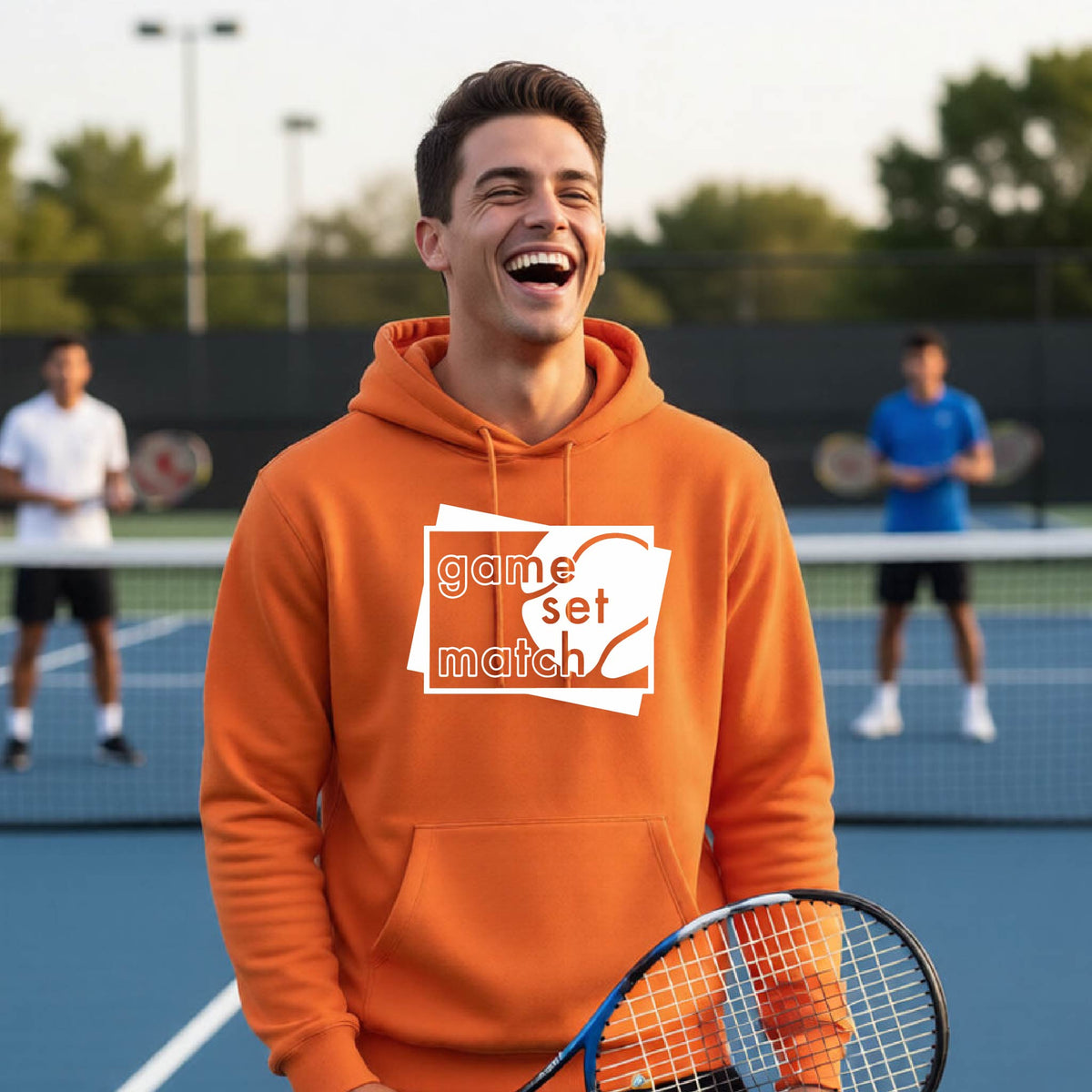 A man on a blue tennis court holds a tennis racket and wears an orange unisex hoodie pullover with a white graphic of two overlapping white and gray squares with the cut out words of ‘GAME SET MATCH” and a tennis ball, printed on the front of the shirt. 