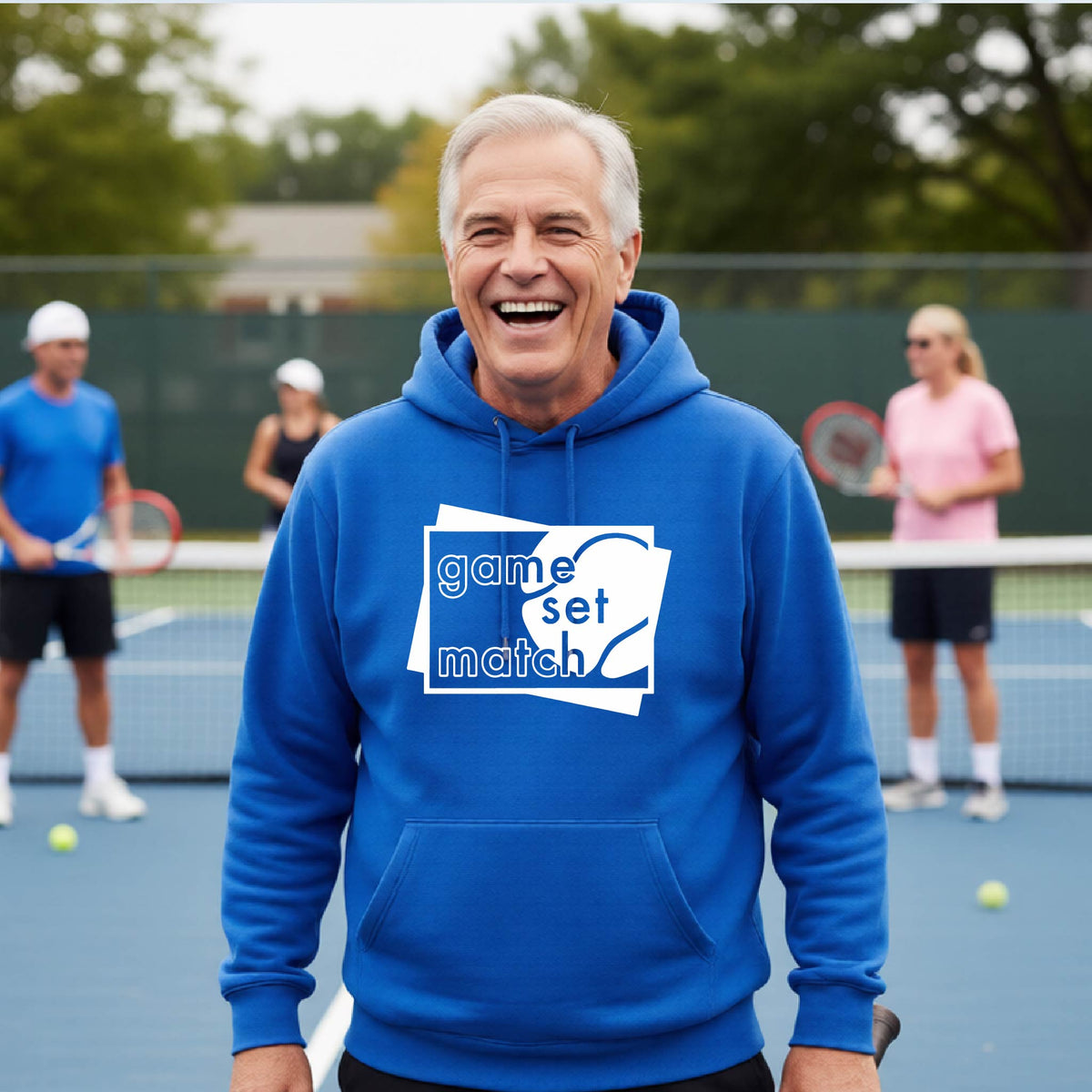 A man on a blue tennis court holds a tennis racket and wears a royal blue unisex hoodie pullover with a white graphic of two overlapping white and gray squares with the cut out words of ‘GAME SET MATCH” and a tennis ball, printed on the front of the shirt. 