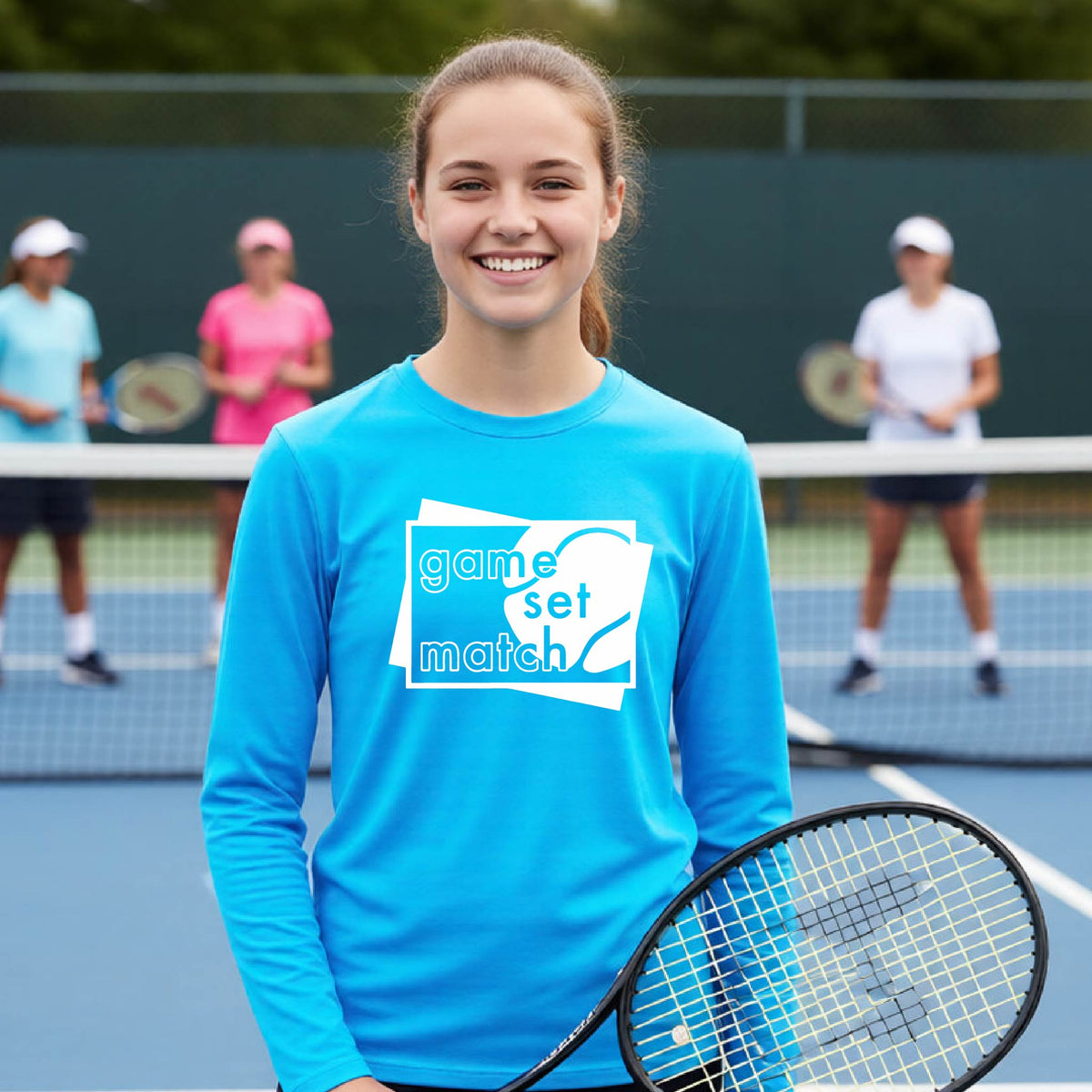 A young person on a blue tennis court holds a tennis racket and wears a bright blue long-sleeve T-shirt with a white graphic of two overlapping white and gray squares with the cut out words of ‘GAME SET MATCH” and a tennis ball, printed on the front of the shirt. 