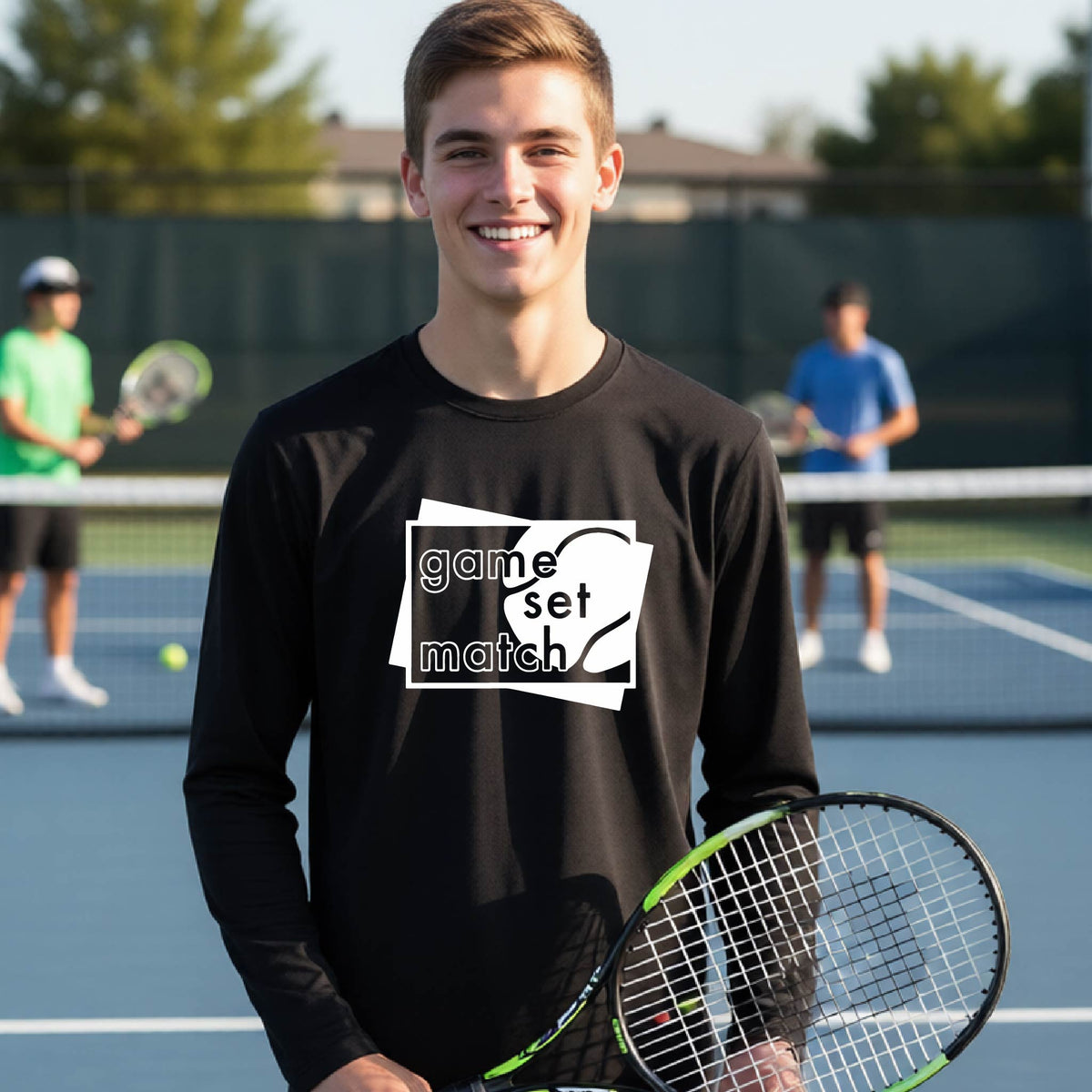 A young person on a blue tennis court holds a tennis racket and wears a black long-sleeve T-shirt with a white graphic of two overlapping white and gray squares with the cut out words of ‘GAME SET MATCH” and a tennis ball, printed on the front of the shirt. 