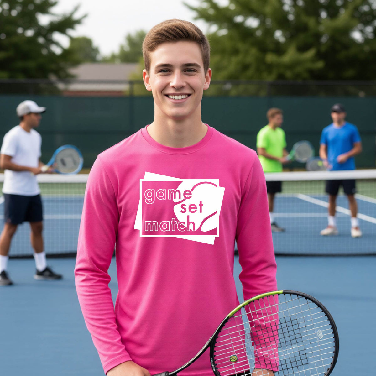 A young person on a blue tennis court holds a tennis racket and wears a pink long-sleeve T-shirt with a white graphic of two overlapping white and gray squares with the cut out words of ‘GAME SET MATCH” and a tennis ball, printed on the front of the shirt. 