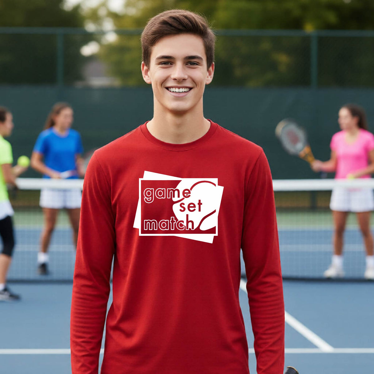 A young person on a blue tennis court holds a tennis racket and wears a red long-sleeve T-shirt with a white graphic of two overlapping white and gray squares with the cut out words of ‘GAME SET MATCH” and a tennis ball, printed on the front of the shirt. 