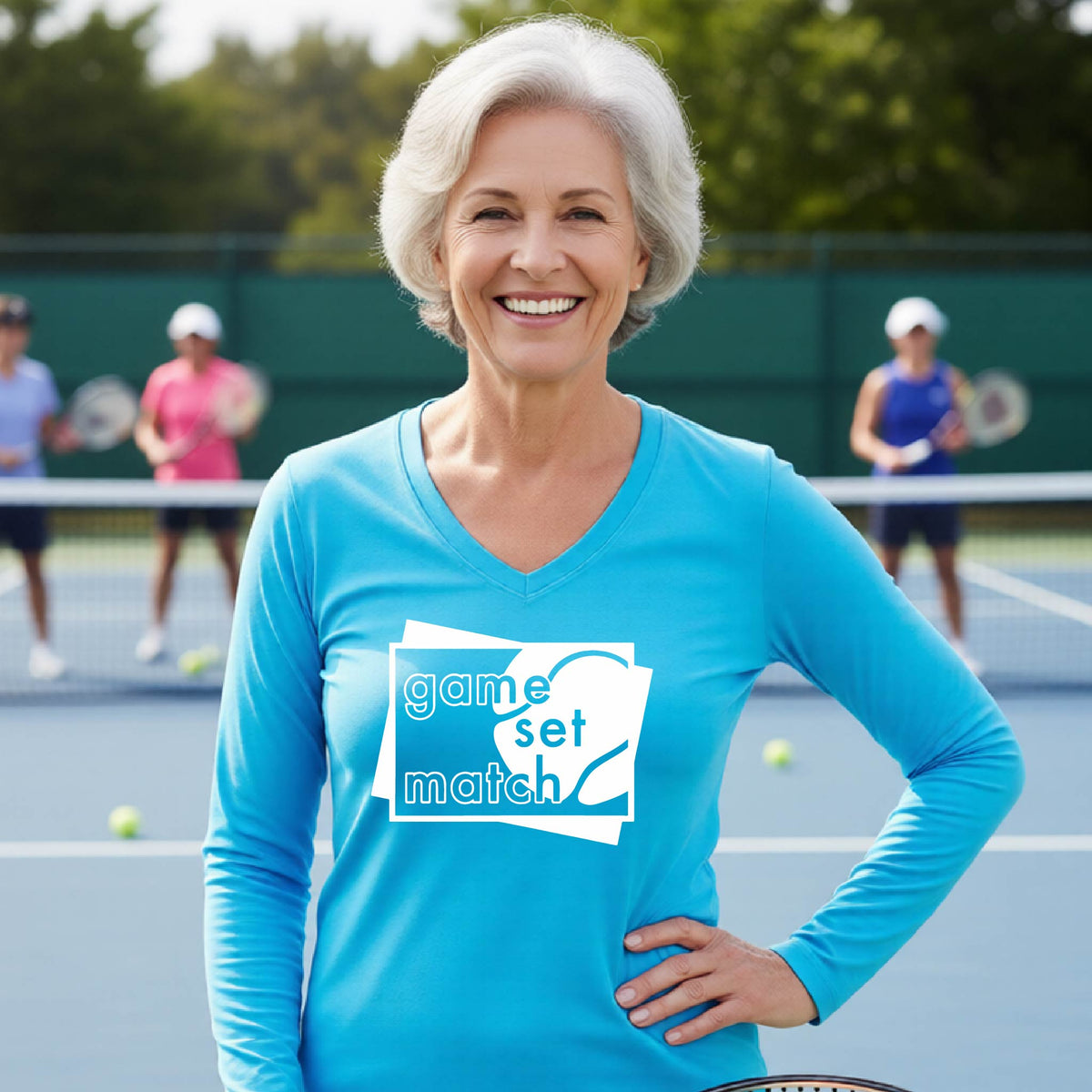 A woman on a blue tennis court holds a tennis racket and wears a bright blue long-sleeve T-shirt with a white graphic of two overlapping white and gray squares with the cut out words of ‘GAME SET MATCH” and a tennis ball, printed on the front of the shirt. 