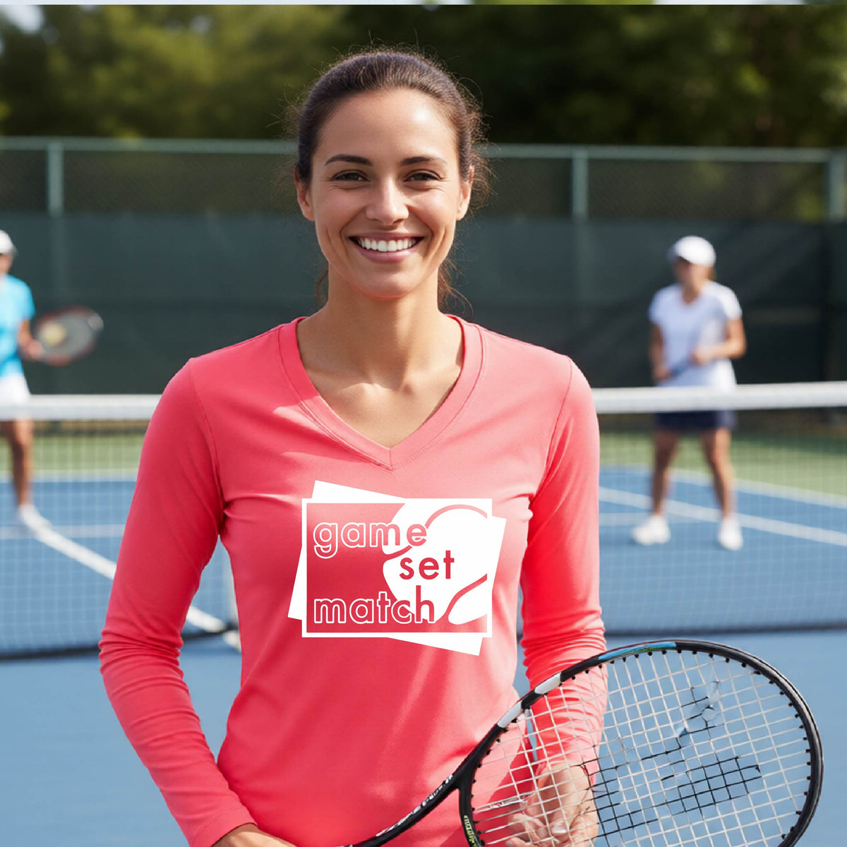 A woman on a blue tennis court holds a tennis racket and wears a coral colored long-sleeve T-shirt with a white graphic of two overlapping white and gray squares with the cut out words of ‘GAME SET MATCH” and a tennis ball, printed on the front of the shirt. 