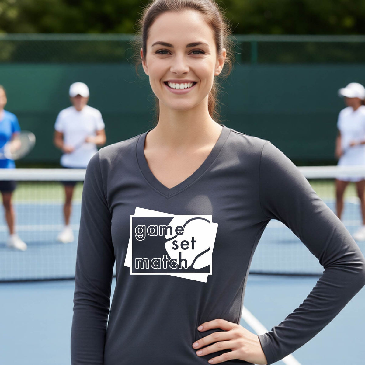 A woman on a blue tennis court holds a tennis racket and wears a dark gray long-sleeve T-shirt with a white graphic of two overlapping white and gray squares with the cut out words of ‘GAME SET MATCH” and a tennis ball, printed on the front of the shirt. 