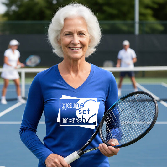 A woman on a blue tennis court holds a tennis racket and wears a royal blue long-sleeve T-shirt with a white graphic of two overlapping white and gray squares with the cut out words of ‘GAME SET MATCH” and a tennis ball, printed on the front of the shirt. 