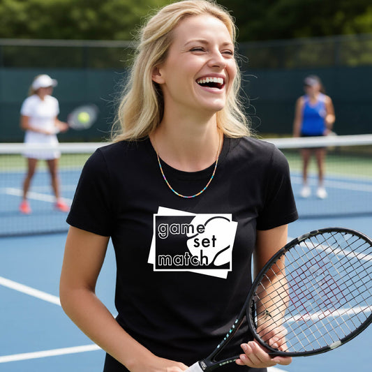 A wpman on a blue tennis court holds a tennis racket and wears a black short-sleeve T-shirt with a white graphic of two overlapping white and gray squares with the cut out words of ‘GAME SET MATCH” and a tennis ball, printed on the front of the shirt. 