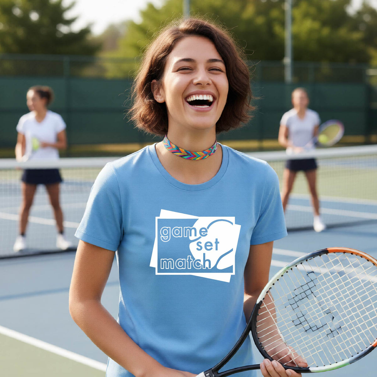 A wpman on a blue tennis court holds a tennis racket and wears a blue short-sleeve T-shirt with a white graphic of two overlapping white and gray squares with the cut out words of ‘GAME SET MATCH” and a tennis ball, printed on the front of the shirt. 