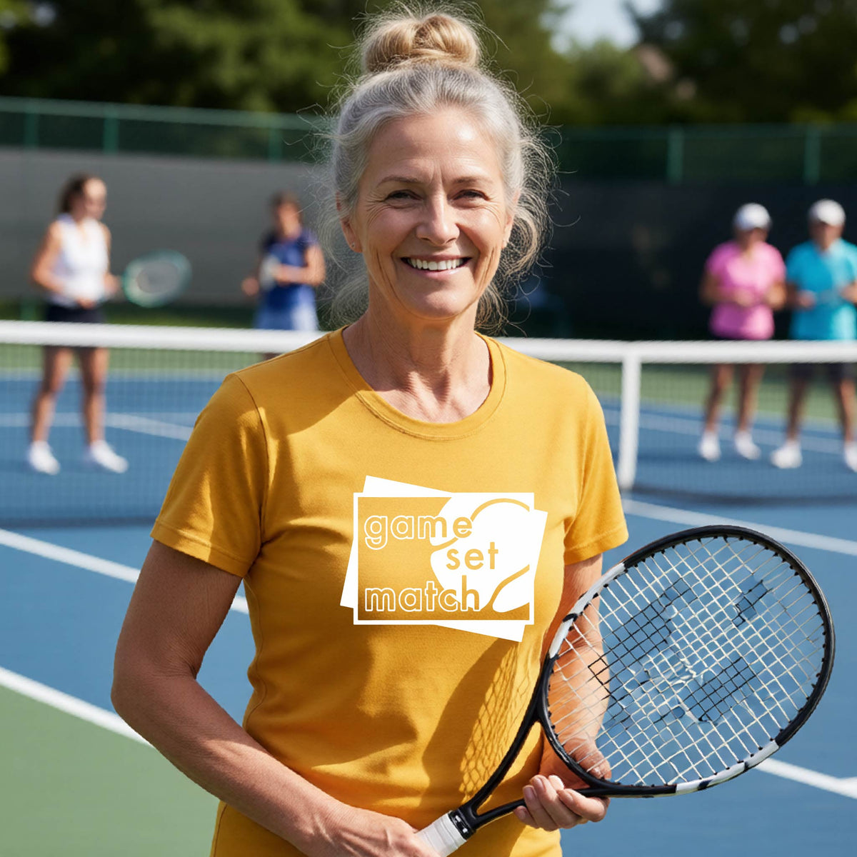 A wpman on a blue tennis court holds a tennis racket and wears a gold short-sleeve T-shirt with a white graphic of two overlapping white and gray squares with the cut out words of ‘GAME SET MATCH” and a tennis ball, printed on the front of the shirt. 
