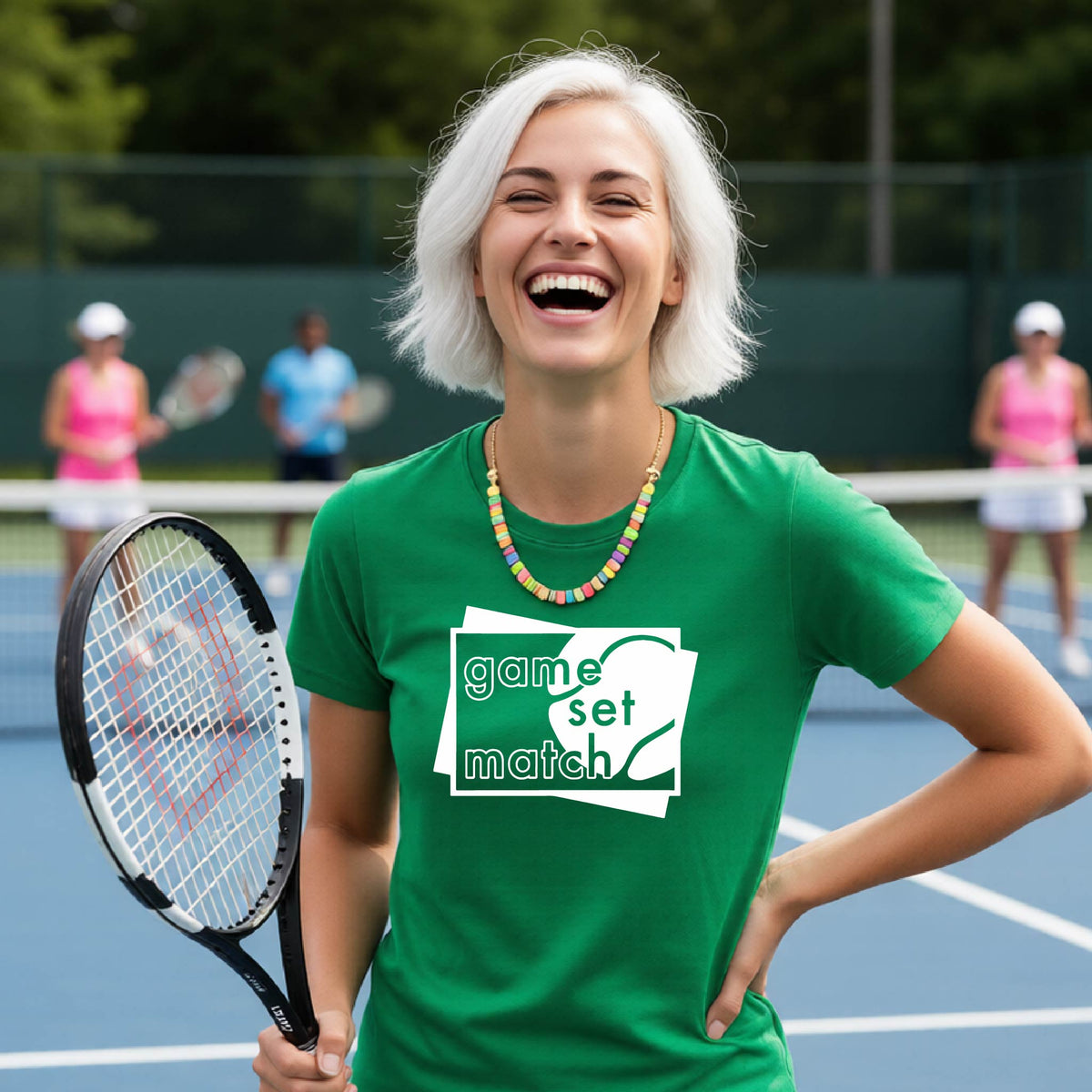 A wpman on a blue tennis court holds a tennis racket and wears a green short-sleeve T-shirt with a white graphic of two overlapping white and gray squares with the cut out words of ‘GAME SET MATCH” and a tennis ball, printed on the front of the shirt. 