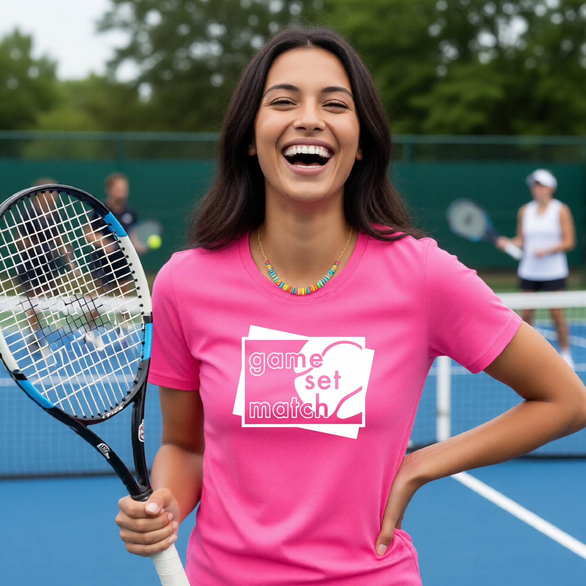 A wpman on a blue tennis court holds a tennis racket and wears a pink short-sleeve T-shirt with a white graphic of two overlapping white and gray squares with the cut out words of ‘GAME SET MATCH” and a tennis ball, printed on the front of the shirt. 