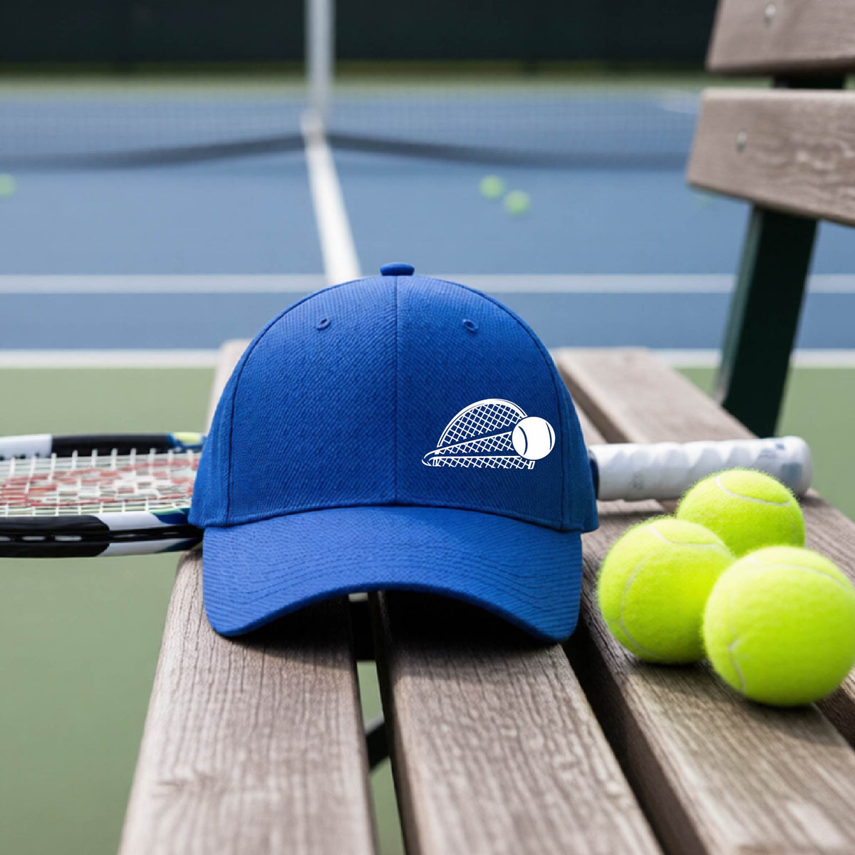 A close-up of a royal blue athletic hat with a white graphic of a half a tennis racquet with a ball streaking over it, on a bench in front of a tennis court.