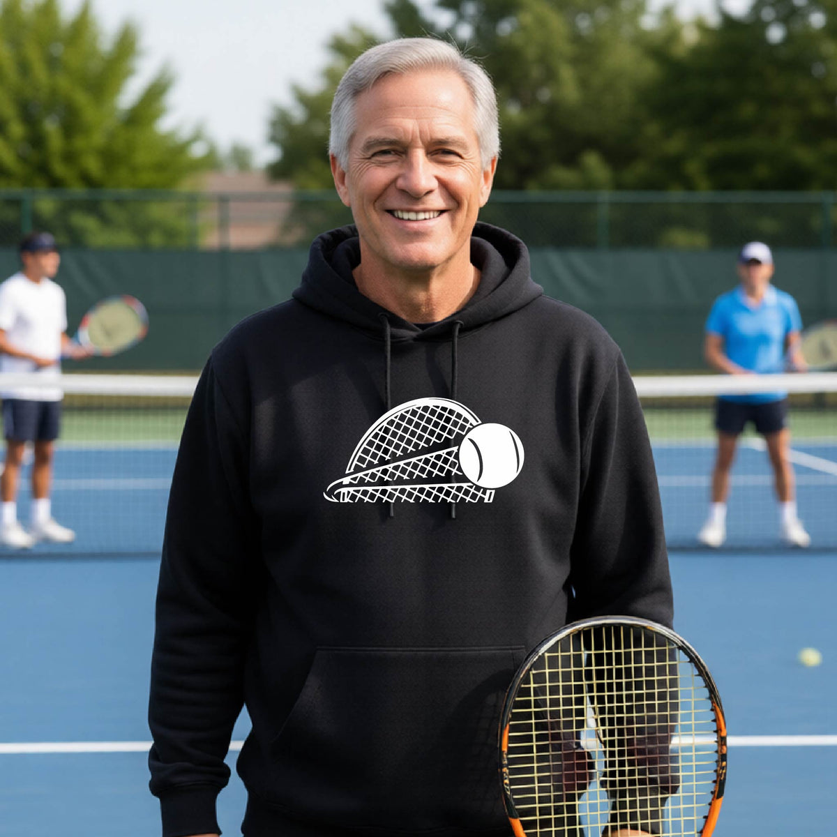 A man on a blue tennis court holds a tennis racket and wears a black unisex hoodie pullover with a white graphic of a half a tennis racquet with a ball streaking over it. 