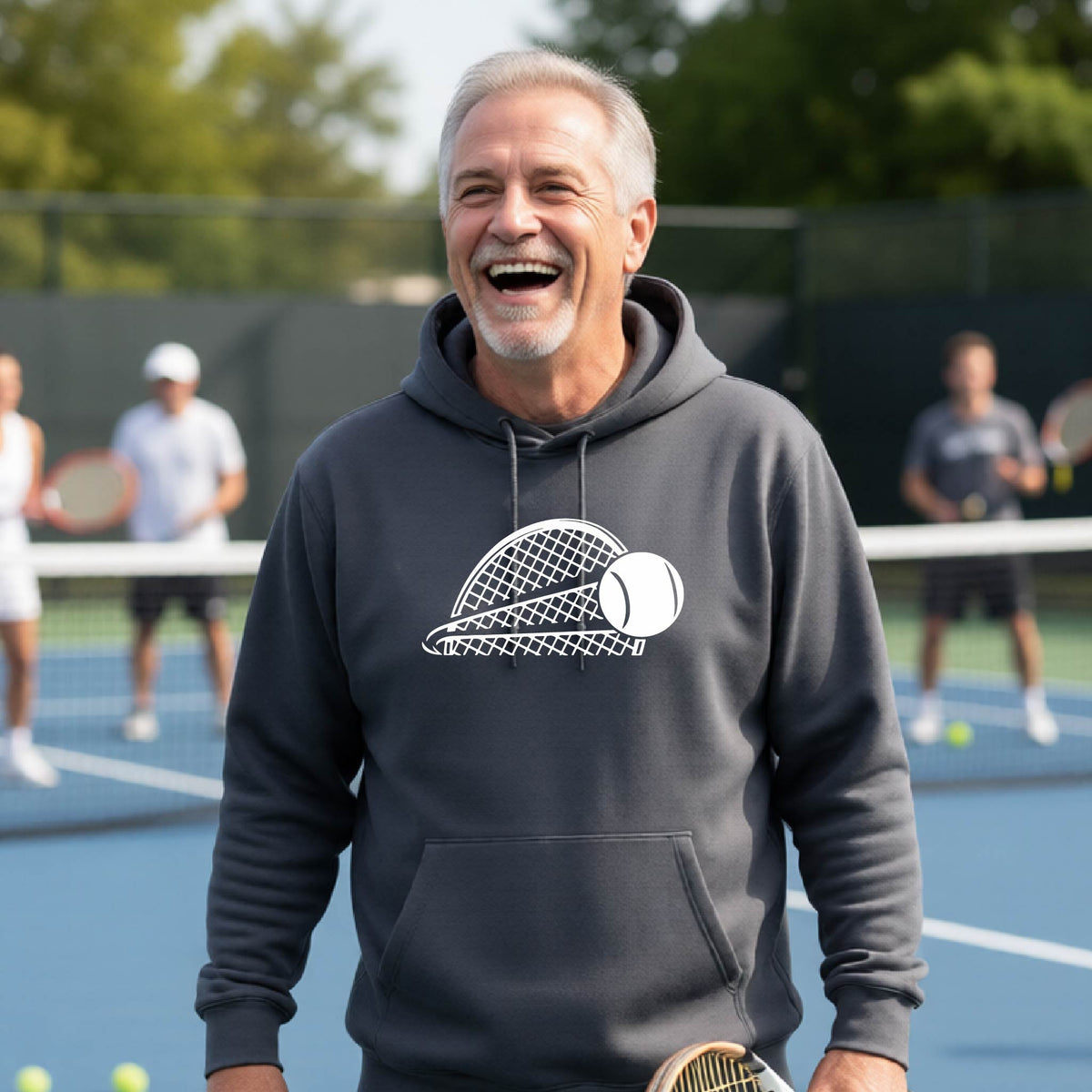 A man on a blue tennis court holds a tennis racket and wears a dark gray unisex hoodie pullover with a white graphic of a half a tennis racquet with a ball streaking over it. 