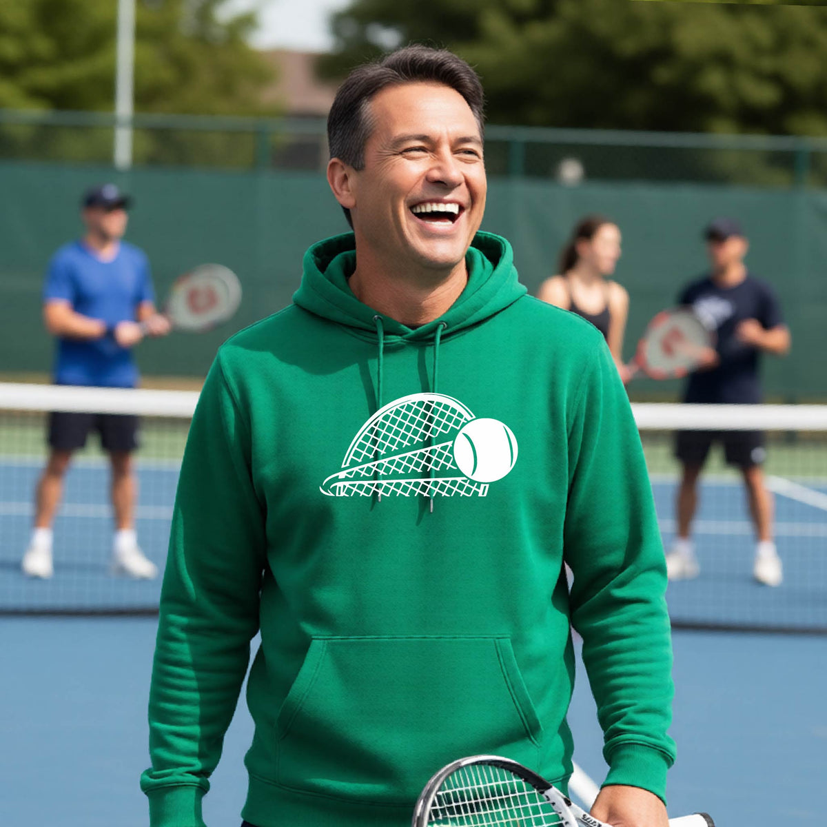 A man on a blue tennis court holds a tennis racket and wears a green unisex hoodie pullover with a white graphic of a half a tennis racquet with a ball streaking over it. 