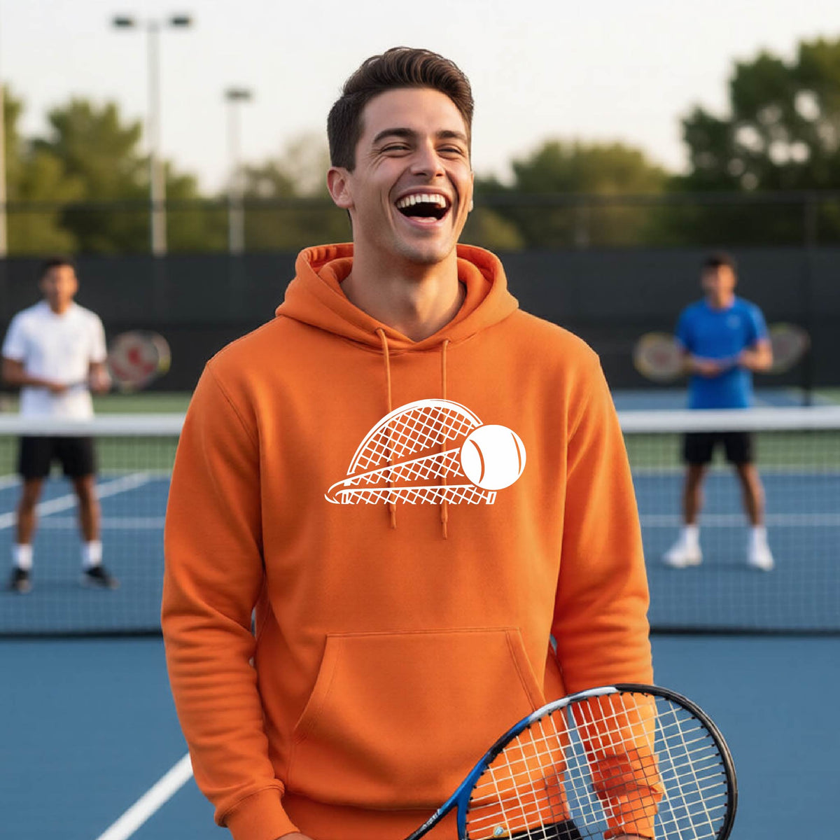 A man on a blue tennis court holds a tennis racket and wears an orange unisex hoodie pullover with a white graphic of a half a tennis racquet with a ball streaking over it. 