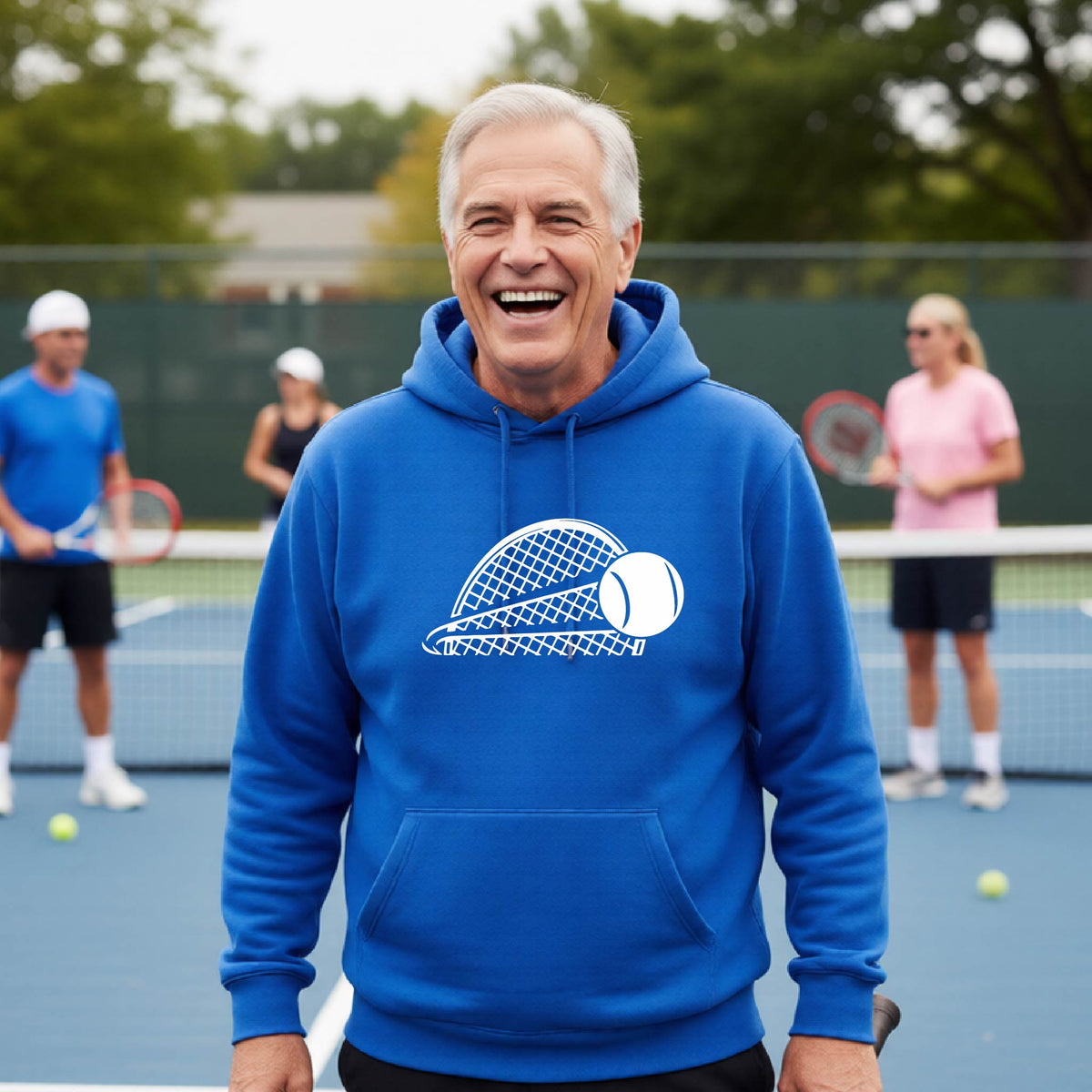A man on a blue tennis court holds a tennis racket and wears a royal blue unisex hoodie pullover with a white graphic of a half a tennis racquet with a ball streaking over it. 