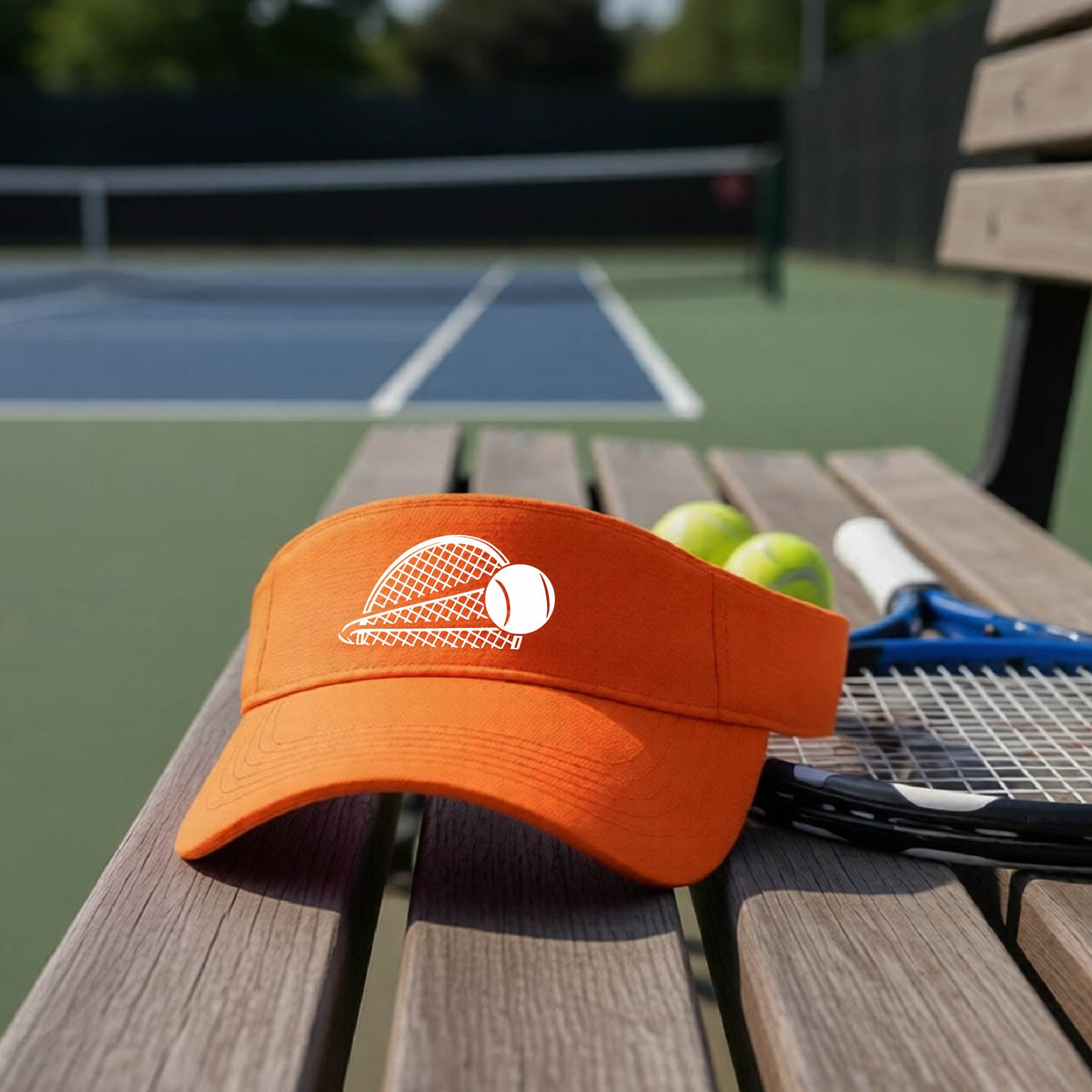 A close-up of an orange athletic visor with a white graphic of a half a tennis racquet with a ball streaking over it, on a bench in front of a tennis court. 