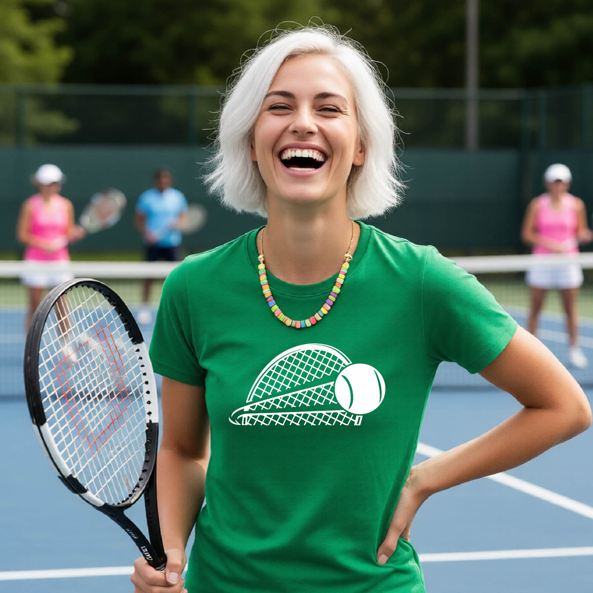 A woman on a blue tennis court holds a tennis racket and wears a green short-sleeve T-shirt with a white graphic of a half a tennis racquet with a ball streaking over it. 