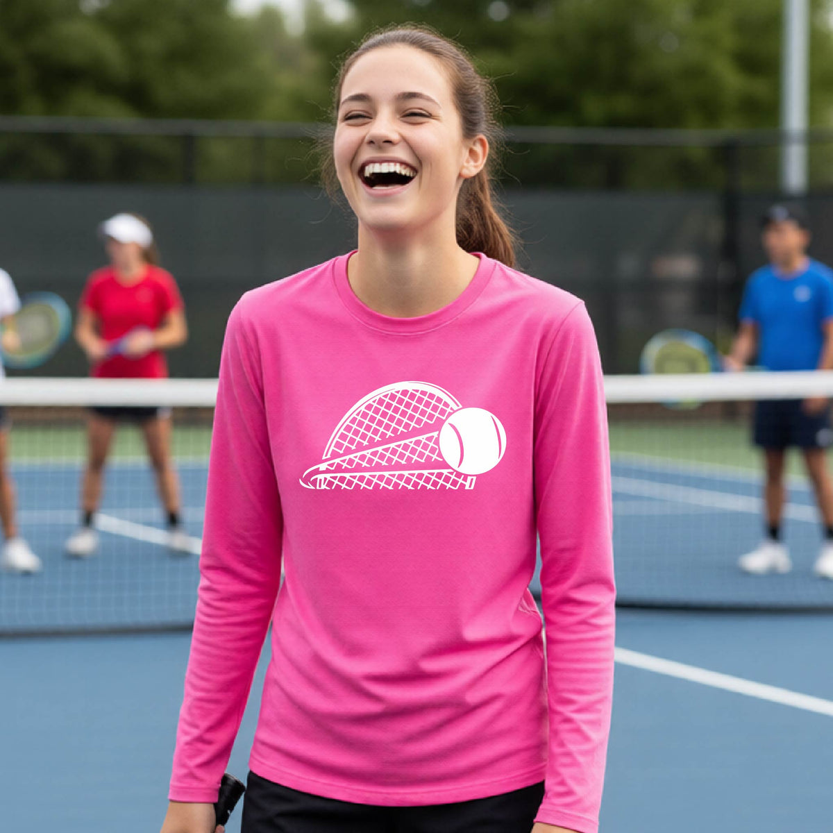 A young person on a blue tennis court holds a tennis racket and wears a pink long-sleeve T-shirt with a white graphic of a half a tennis racquet with a ball streaking over it. 