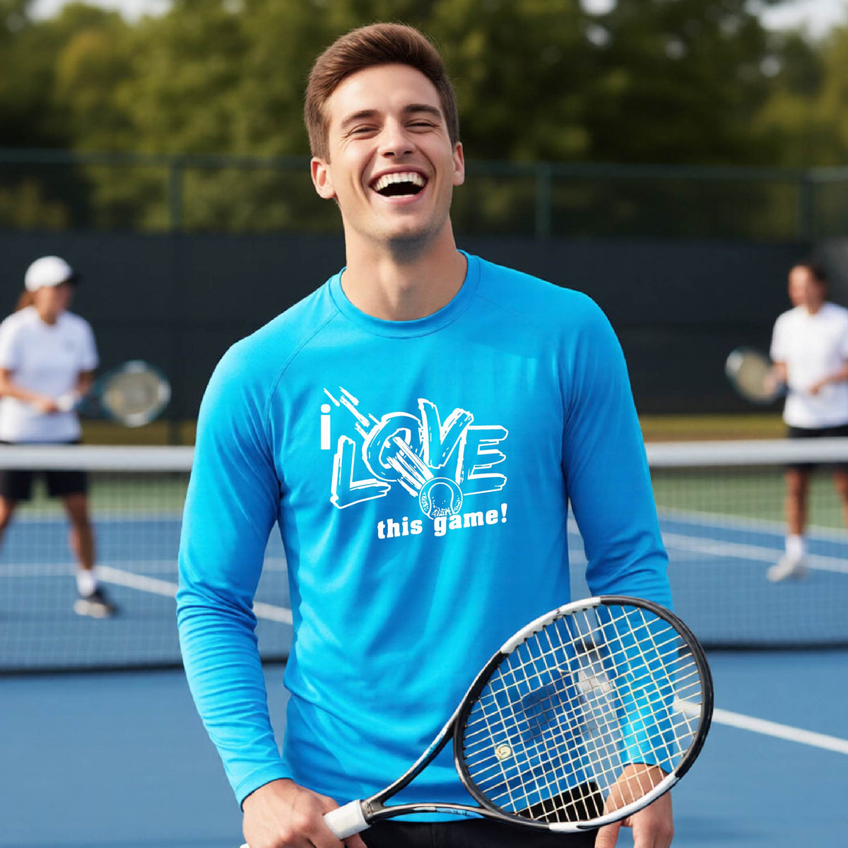 A man on a tennis court wearing a bright blue long-sleeve athletic shirt with the text 'i LOVE this game!' printed on the front with a white tennis ball, holding a tennis racket. Two other players are blurred in the background on the court.