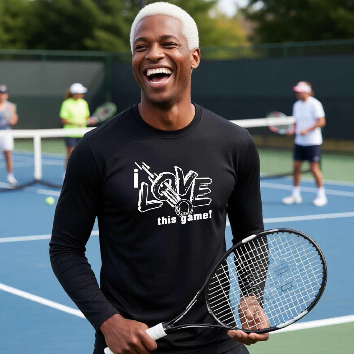 A man with platinum blonde hair laughing on a tennis court, wearing a black long-sleeve t-shirt with a white graphic that reads 'i LOVE this game!' and a tennis ball. He is holding a tennis racket, with other players and the court visible in the background.
