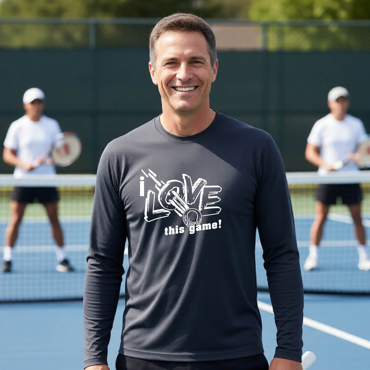 A smiling man wearing a dark gray long-sleeve t-shirt with a white graphic that reads 'i LOVE this game!' and a whit tennis icon, stands on a tennis court. Two blurred players are visible in the background behind a net.