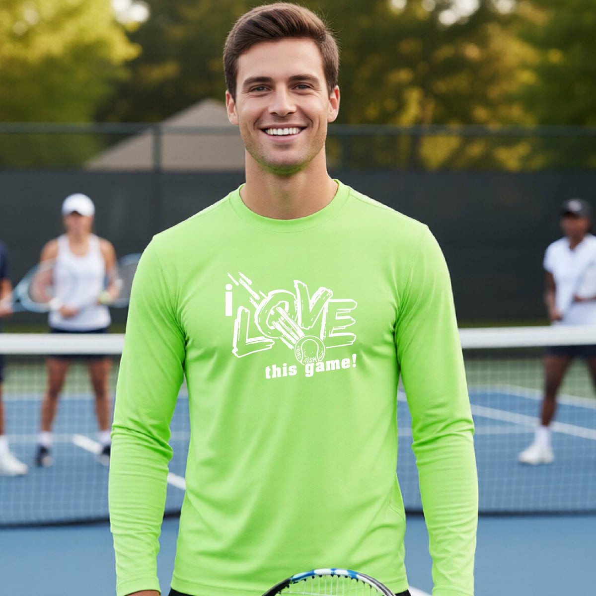 A smiling man wearing a lime green long-sleeve t-shirt with a white graphic that reads 'i LOVE this game!' and a whit tennis icon, stands on a tennis court. Two blurred players are visible in the background behind a net.