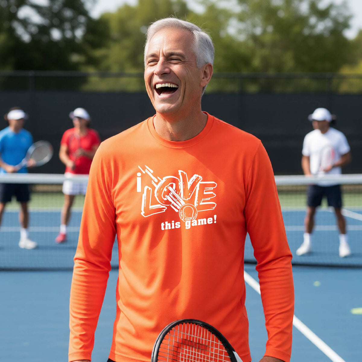 A smiling older man on a tennis court wearing a bright orange long-sleeve shirt with a white graphic that reads 'i LOVE this game!' and a white tennis ball. Other tennis players are blurred in the background on a blue court.