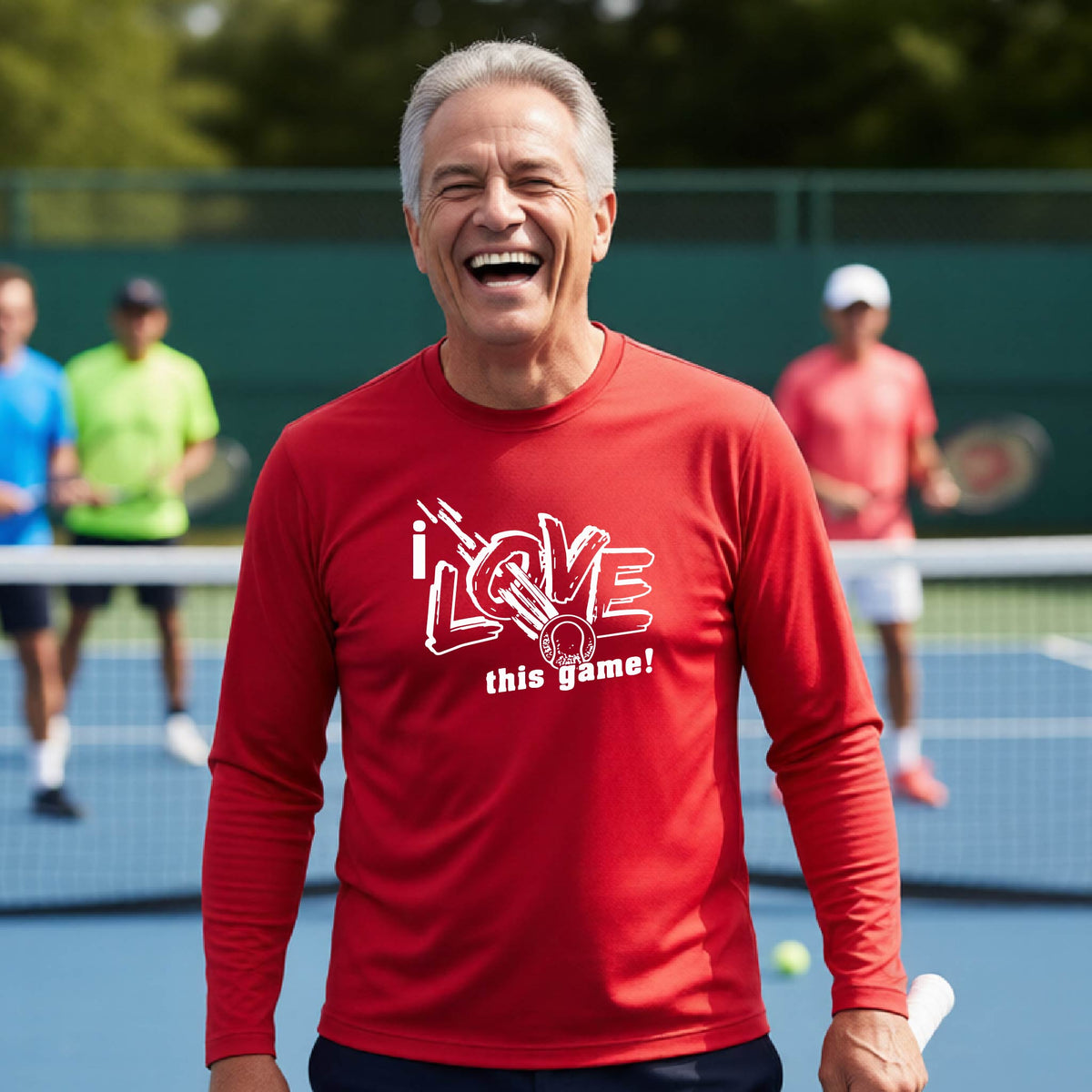 A smiling man wearing a red long-sleeve t-shirt with a white graphic that reads 'i LOVE this game!' and a whit tennis icon, stands on a tennis court. Two blurred players are visible in the background behind a net.