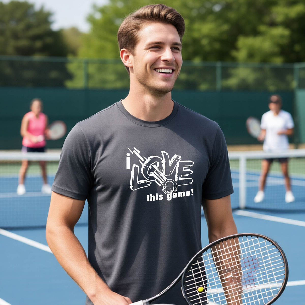 A smiling man on a tennis court wearing a dark gray t-shirt with a white graphic that reads 'i LOVE this game!'. He is holding a tennis racket, with other players and a green fence blurred in the background.