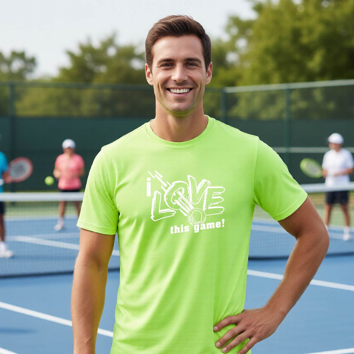 A smiling man wearing a bright neon green t-shirt with a white graphic that reads 'i LOVE this game!' stands on a tennis court. The background shows a blurred tennis court with other players and trees.