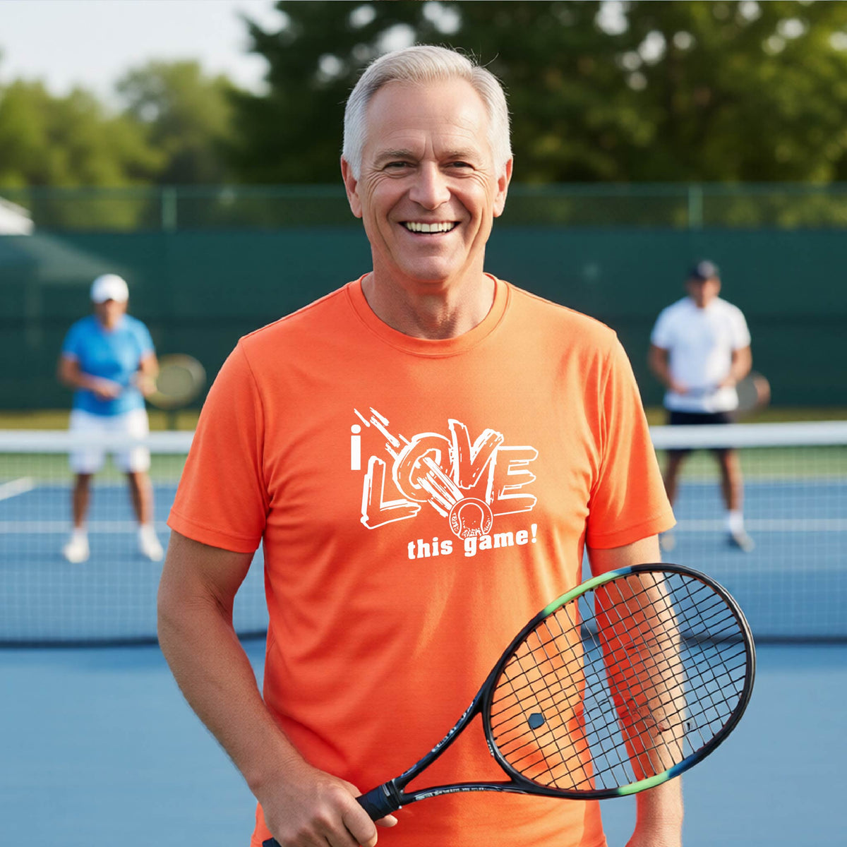 A smiling older man on a tennis court wearing a bright orange T-shirt with the text 'i LOVE this game!' and holding a tennis racket. Two other players are blurred in the background near the net.