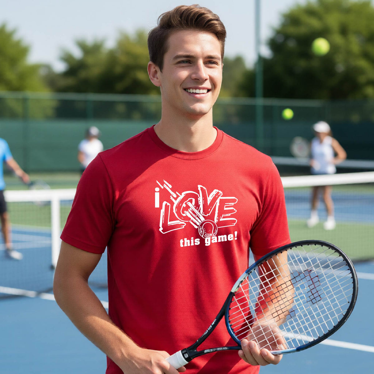 A smiling man on a tennis court wearing a red graphic T-shirt that reads 'i LOVE this game!' in white, holding a tennis racket. Other tennis players and a green fence are blurred in the background.