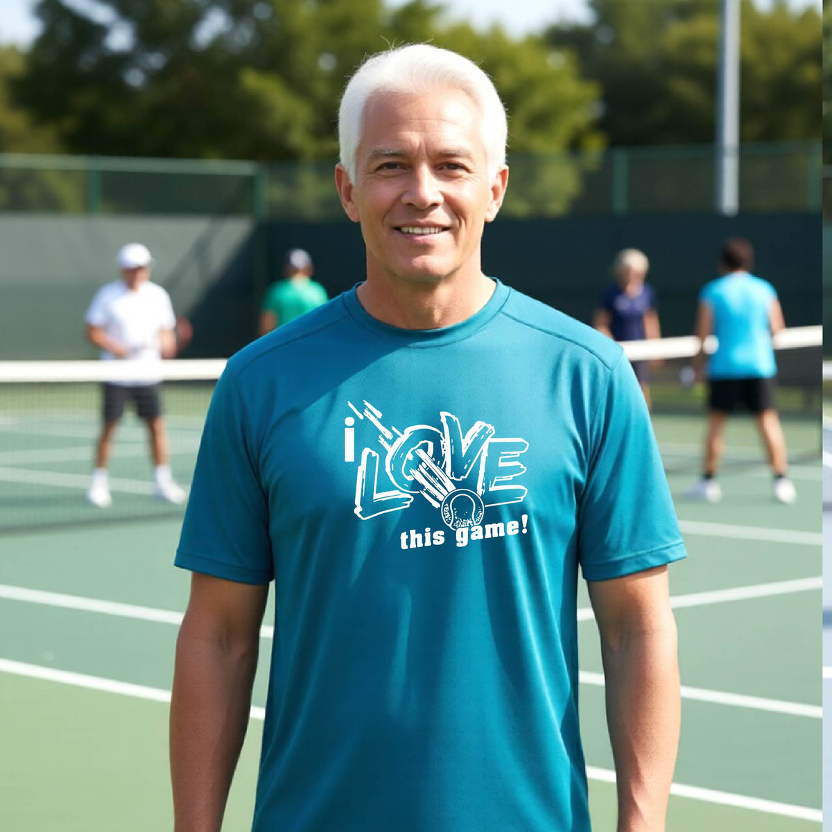 A smiling man with white hair stands on a tennis court wearing a teal blue t-shirt with a white graphic that reads 'i LOVE this game!'. Other players are visible in the background on the court.