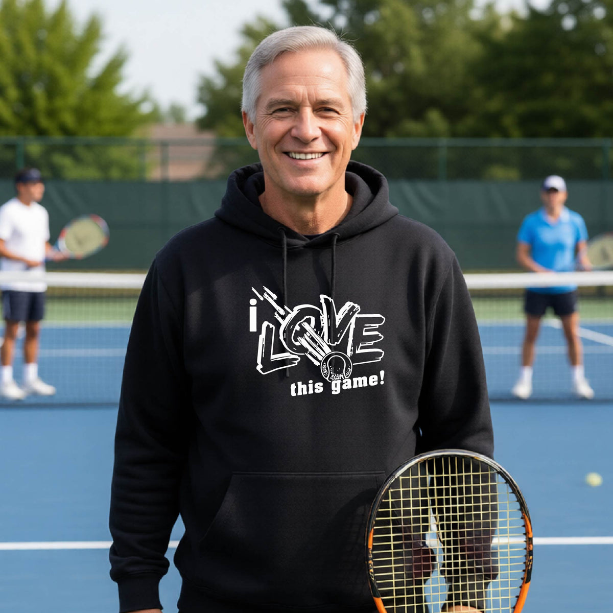 A smiling man wearing a black hoodie with a white graphic that reads 'i LOVE this game!' stands on a blue tennis court holding a tennis racket. Two other players are visible in the background.
