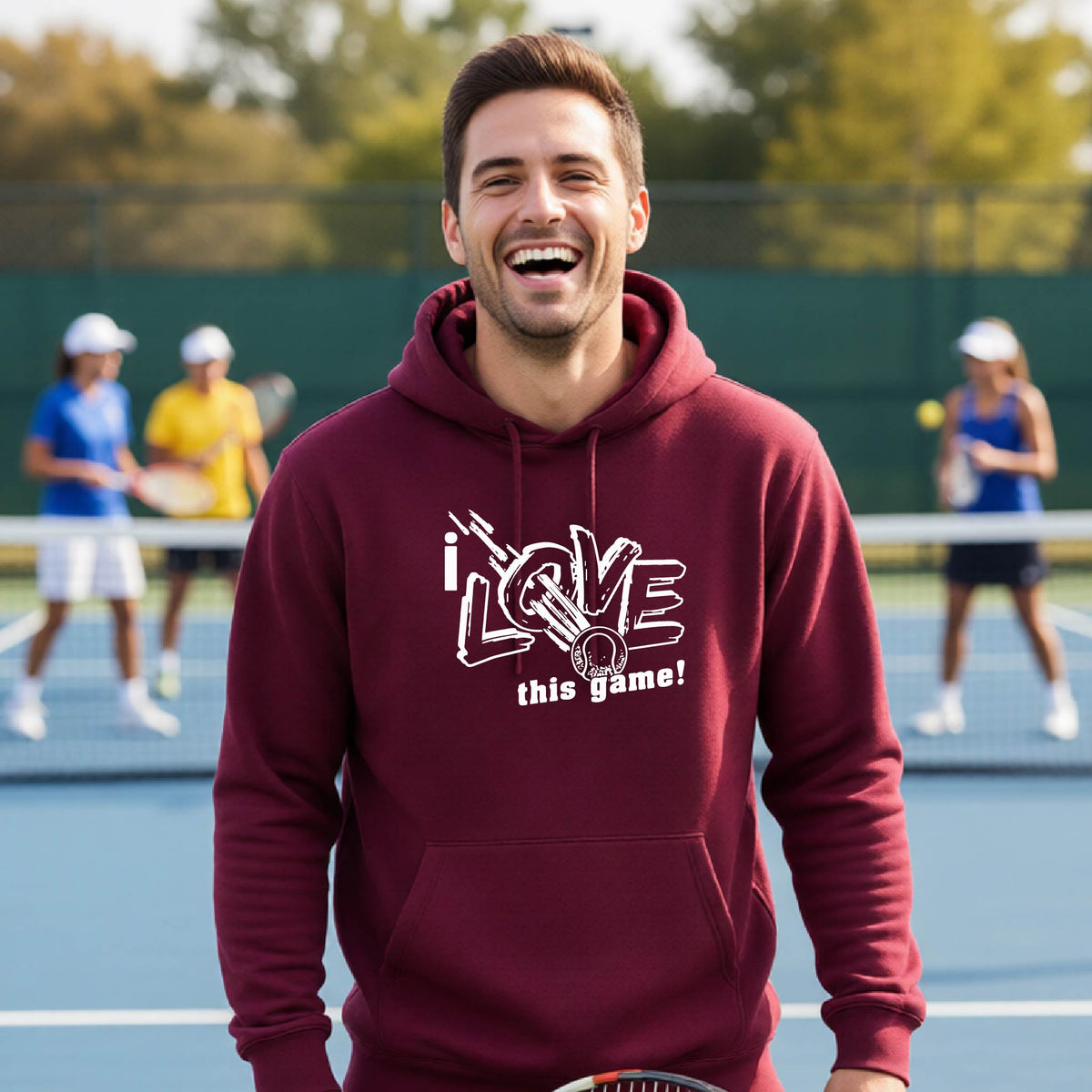 A man wearing a maroon hoodie with a white graphic that reads 'i LOVE this game!' stands on a tennis court, with blurred players in the background.