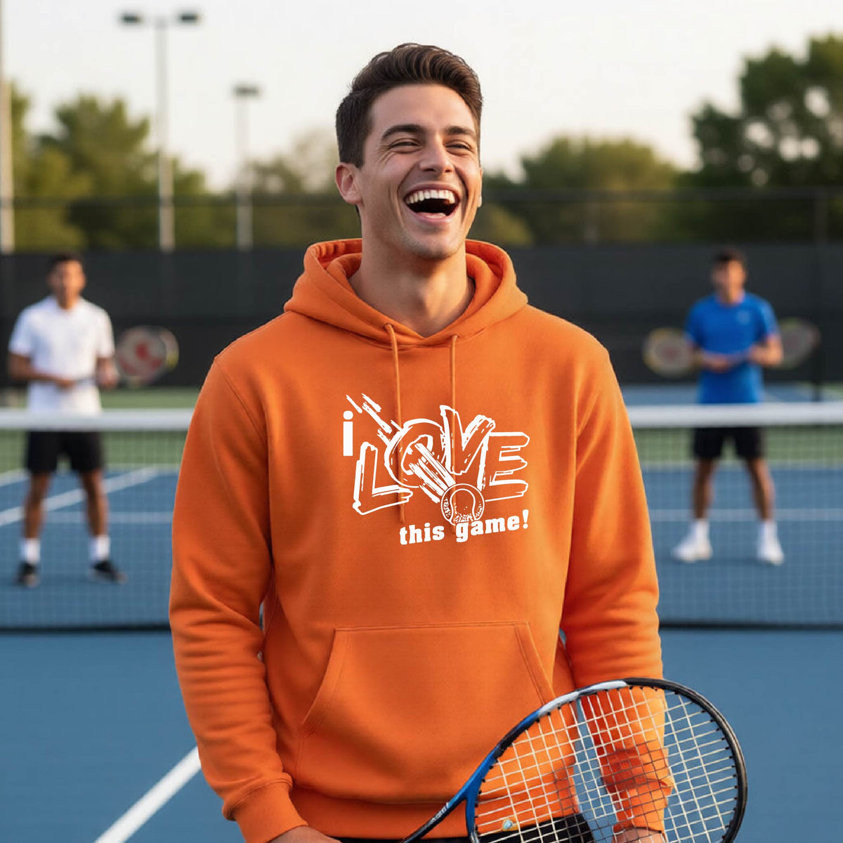 A man laughing on a tennis court wearing an orange hoodie with a white graphic that reads 'i LOVE this game!'. Two blurred tennis players are visible in the background on the court.