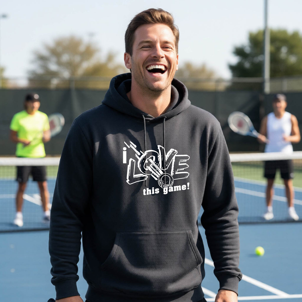 A man laughing on an outdoor tennis court wearing a dark gray hoodie with a white graphic that reads 'i LOVE this game!'. Two other tennis players are visible in the background behind the net.