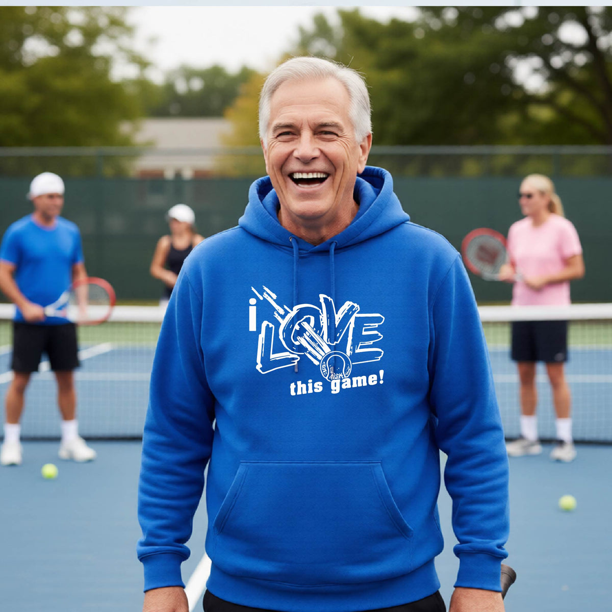 A smiling older man wearing a bright blue hoodie with a white graphic that reads 'I LOVE this game!' stands on a tennis court, with other players blurred in the background.