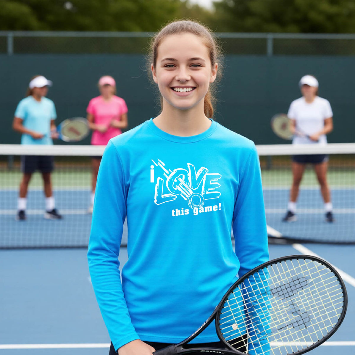 A smiling young woman stands on a tennis court holding a racket, wearing a bright blue long-sleeve shirt with the graphic text 'I LOVE this game!'. Two other players are visible in the background behind the net.