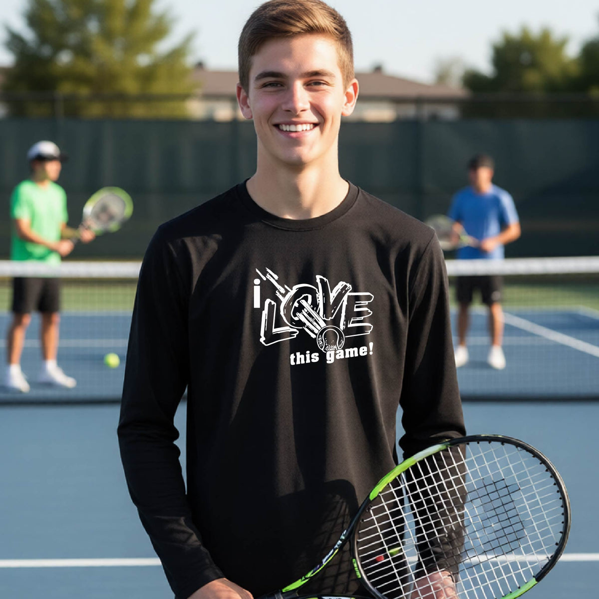 A smiling young man on a tennis court wearing a black long-sleeve t-shirt with a white graphic that reads 'I LOVE this game!'. He is holding a tennis racket, with other players and tennis courts blurred in the background.